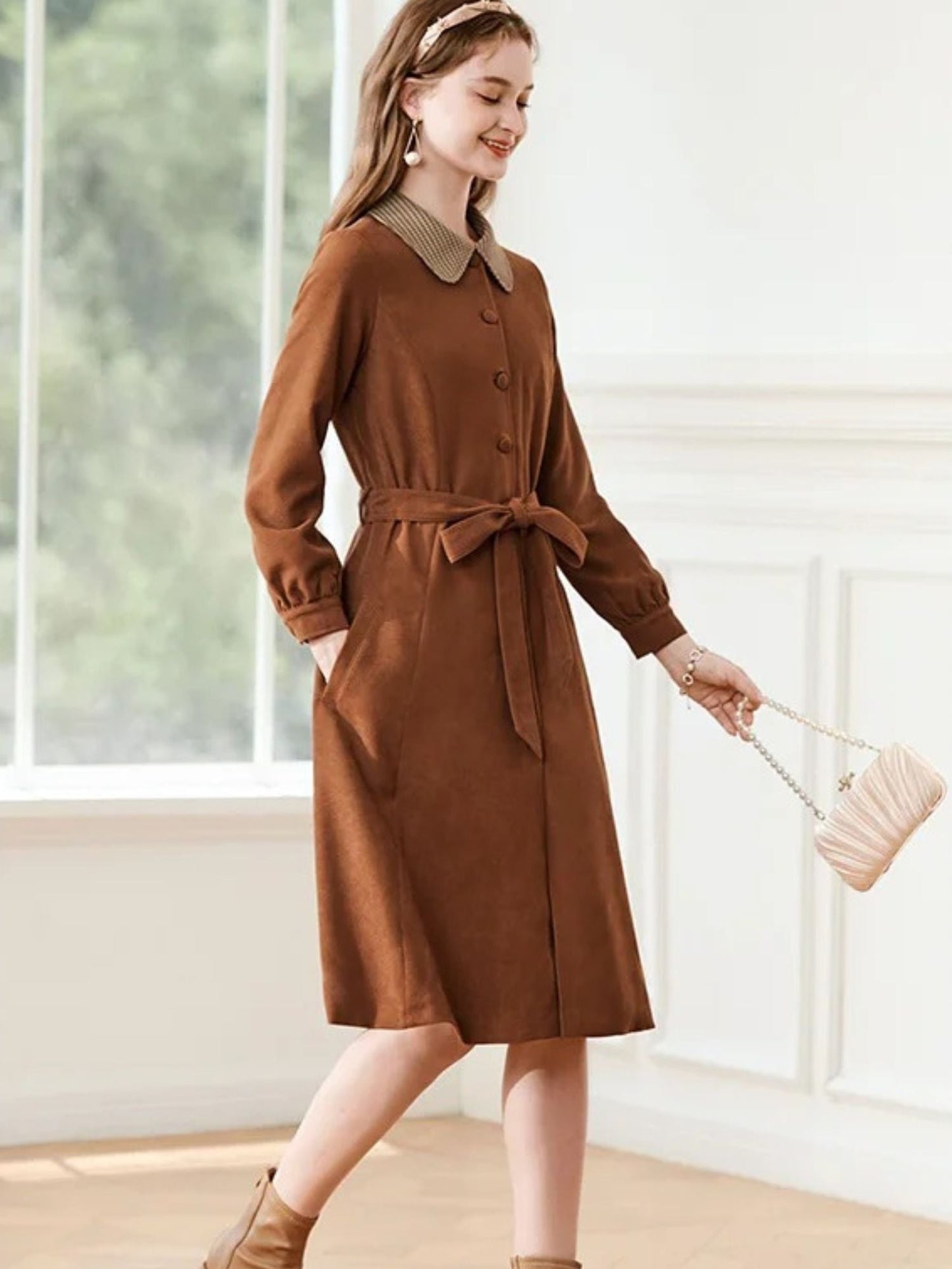Woman walking in brown dress with tie waist and patterned collar, holding a beige clutch.