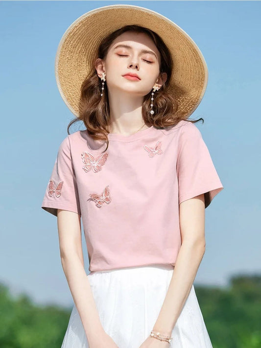 Woman smiling in pink tee with butterfly details, holding her hat in the sun.