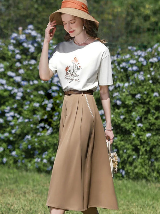 Side view of woman in tan skirt and white graphic shirt, holding her hat while walking.
