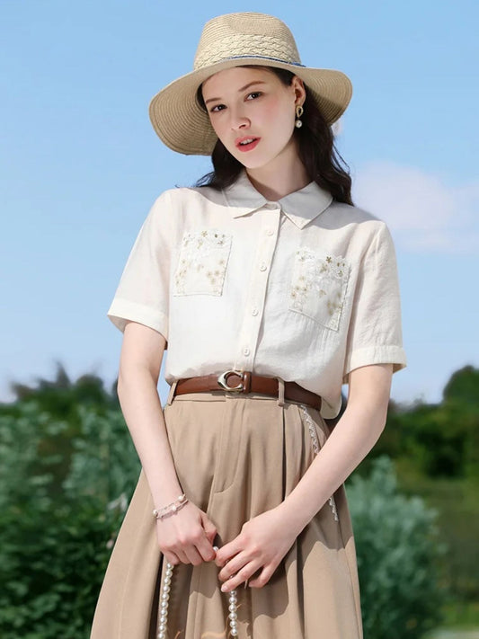 Woman in white floral blouse and pleated skirt posing in a garden with hands folded.