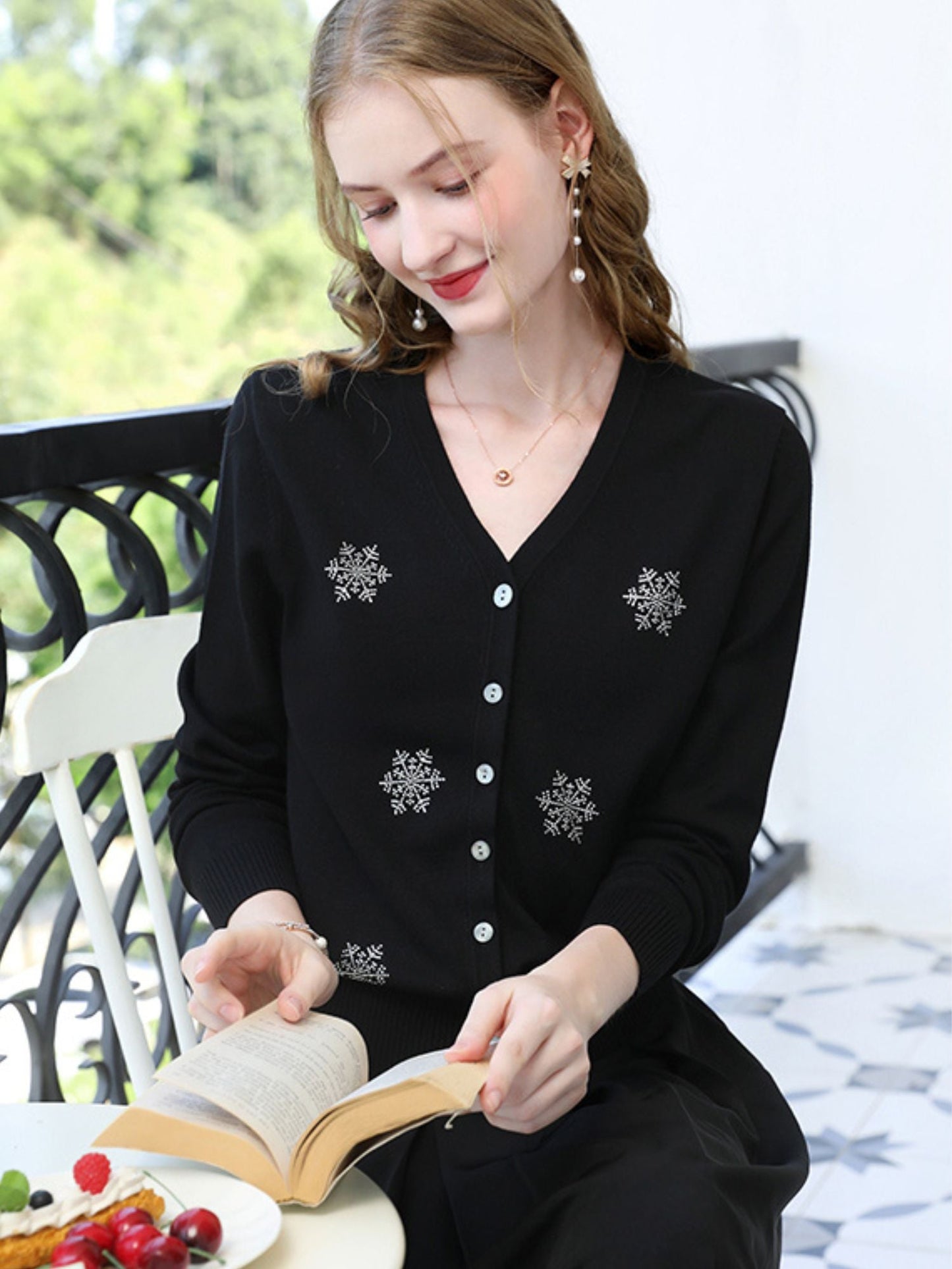 Woman sitting at a table in black snowflake cardigan, smiling with a book and fruit nearby.