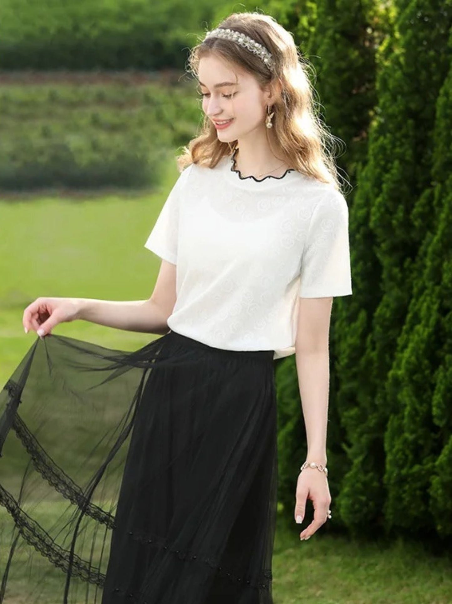 Side view of woman in white short-sleeve top with black trim, smiling near trees.