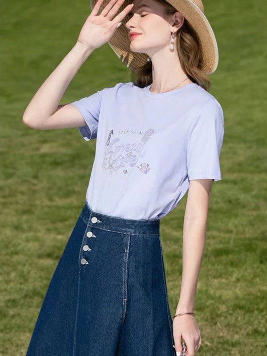 Side view of woman in tee and denim midi skirt, touching her hat and smiling in the sun.