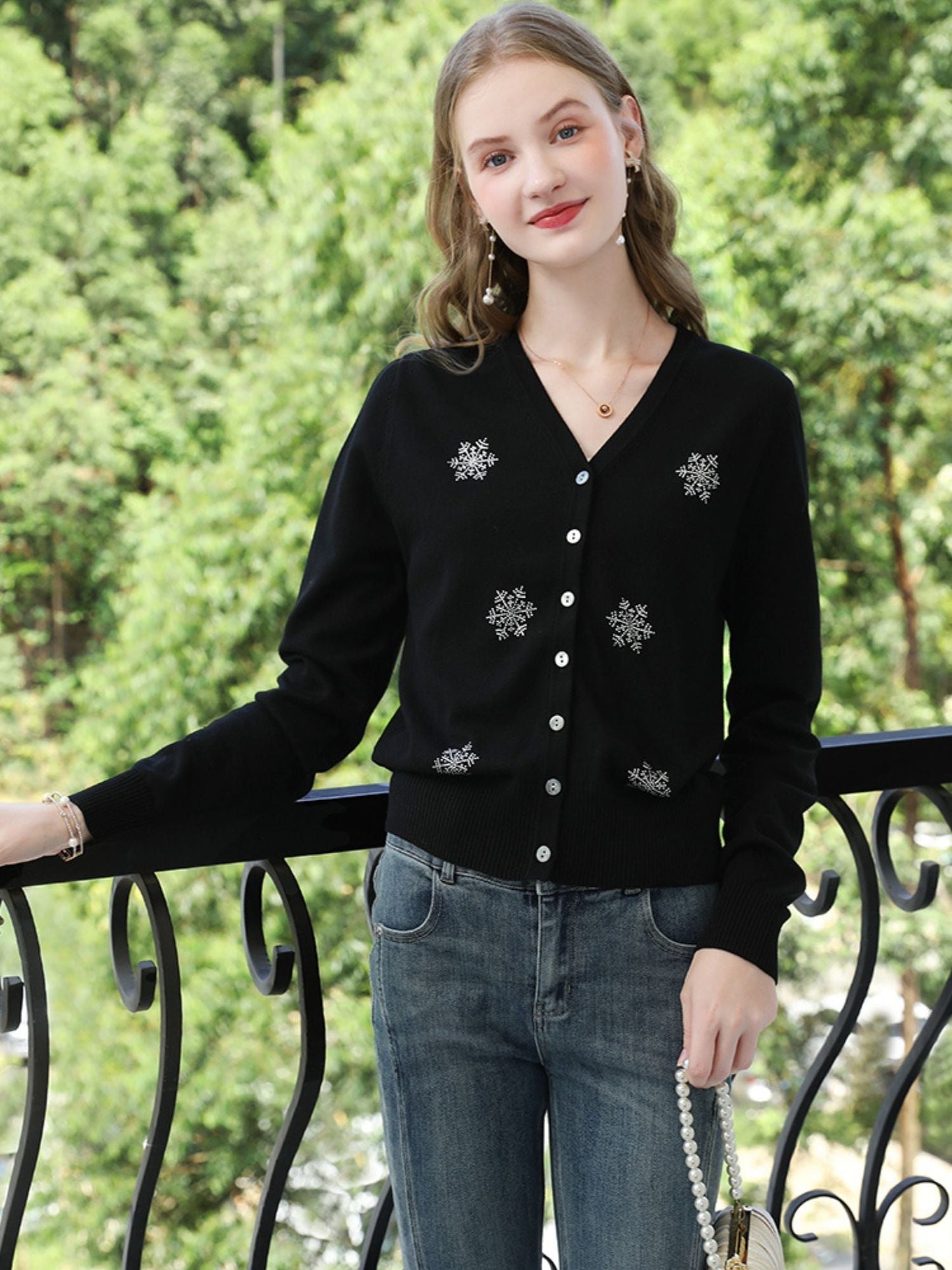 Woman in black embroidered cardigan and jeans, smiling outdoors near trees.