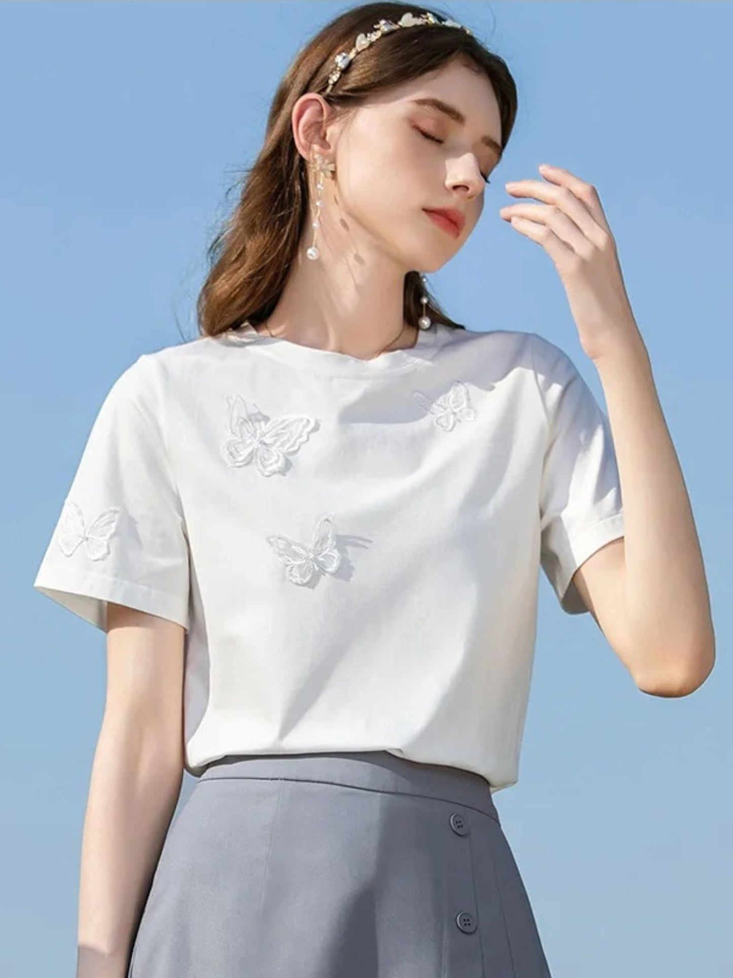 Woman in white embroidered tee and gray skirt, touching her hair.