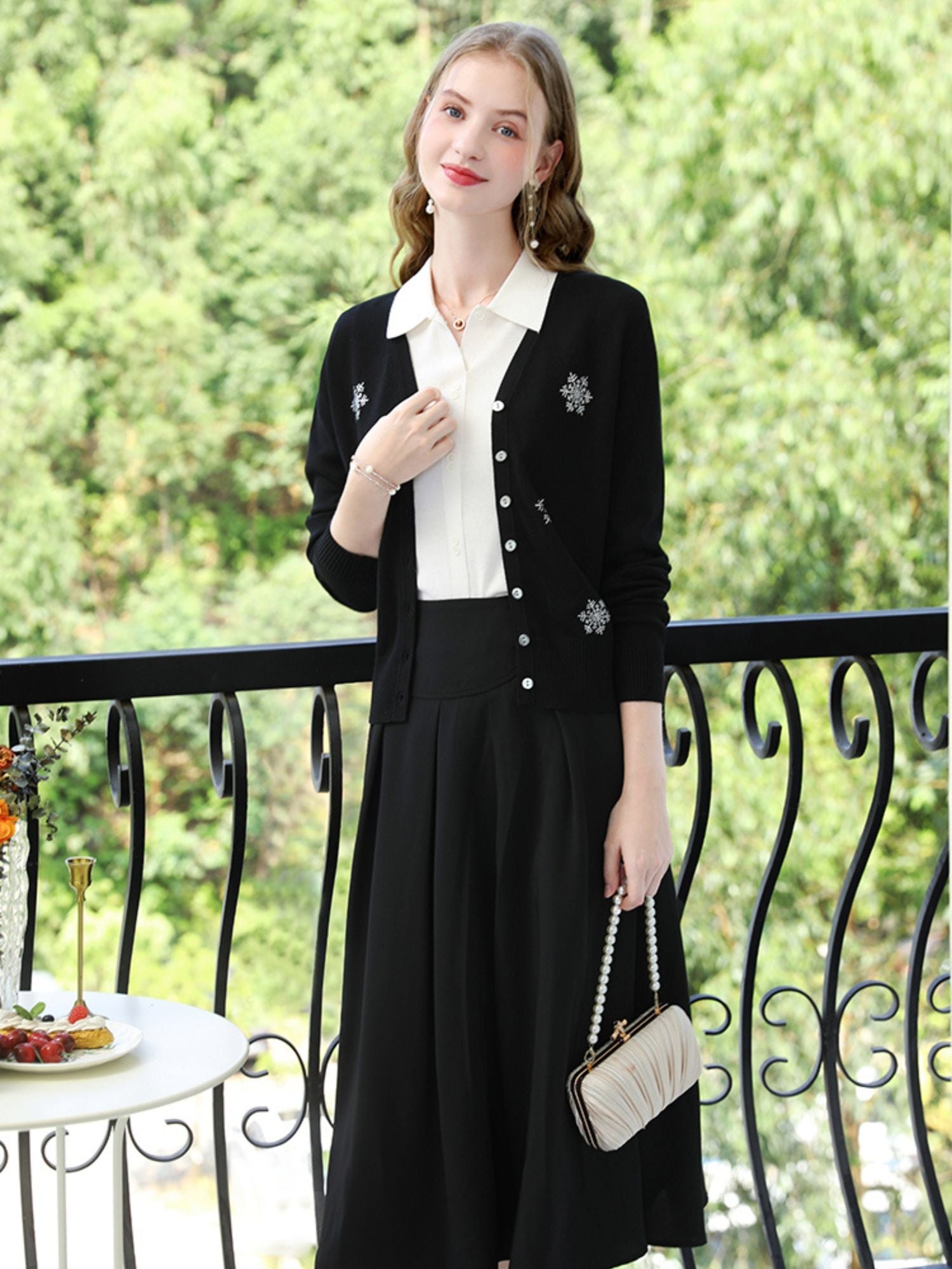Woman in black cardigan with snowflake embroidery over a white top, holding a small quilted purse.