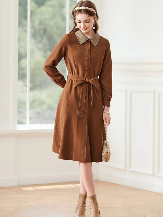 Woman in brown belted dress with houndstooth collar, holding a woven bag and looking down.