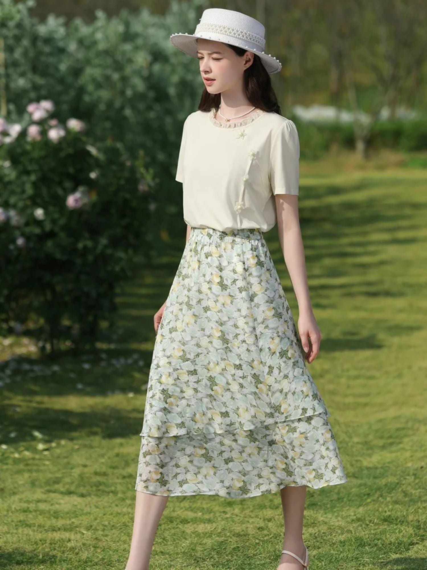 Woman in cream blouse and floral midi skirt with tiered layers, wearing a white hat outdoors.