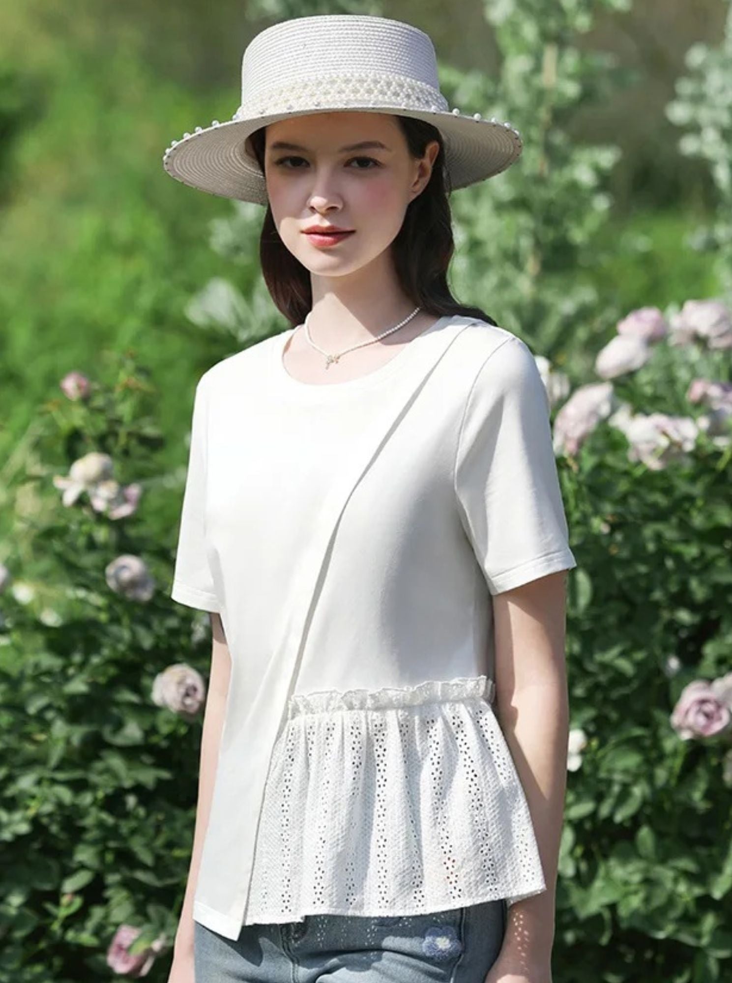 Woman wearing a white eyelet peplum top and straw hat, standing in front of blooming bushes.