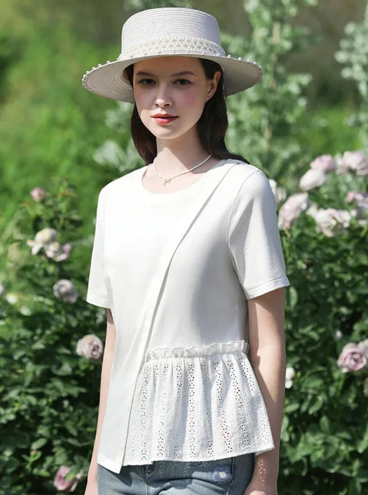 Woman wearing a white eyelet peplum top and straw hat, standing in front of blooming bushes.