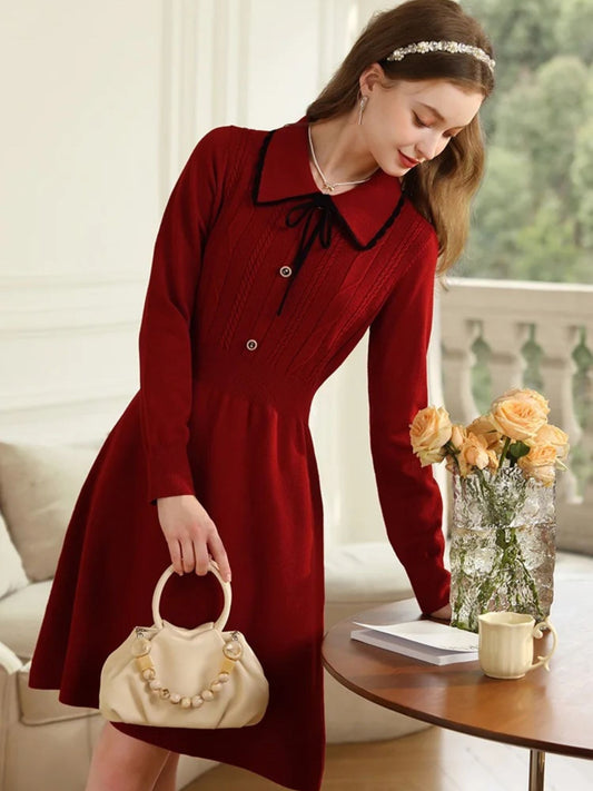 Woman in red A-line knit dress with black collar holding a beige handbag beside a table with yellow roses.