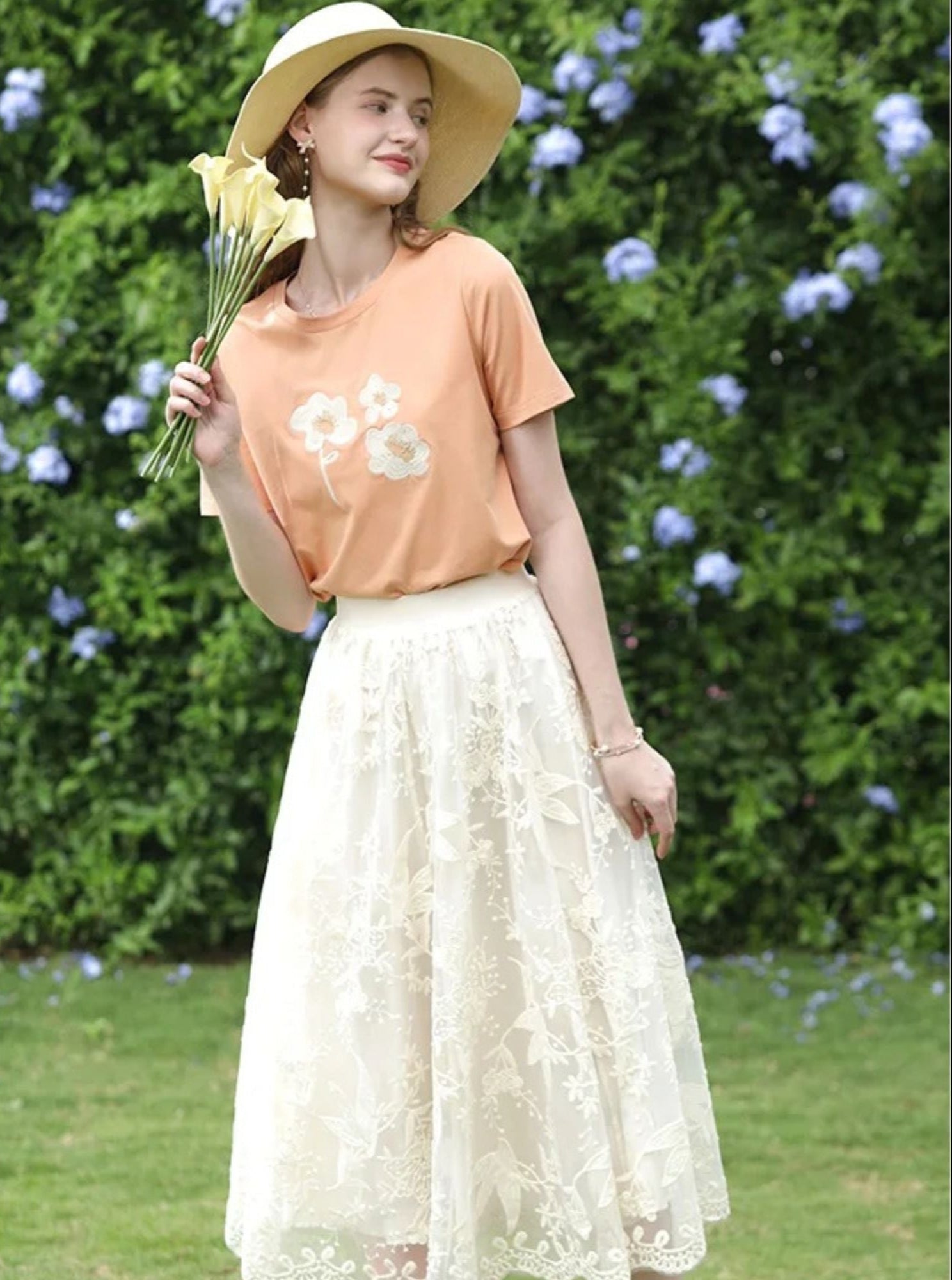 Woman in orange floral tee and lace midi skirt holding yellow flowers and smiling in a sunhat.
