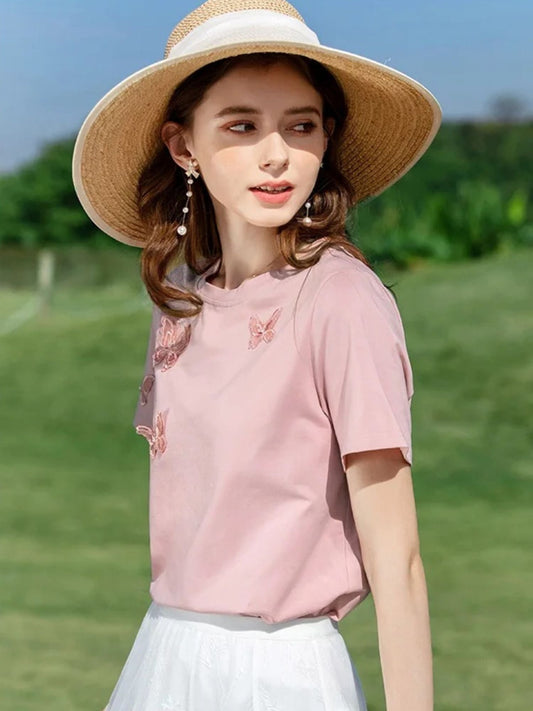 Woman in pink embroidered butterfly tee and white skirt, wearing a straw hat outdoors.