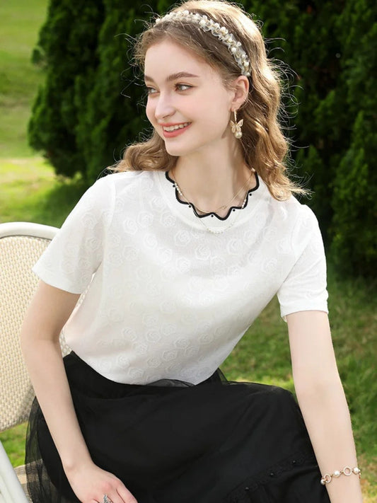 Woman sitting in white lace top with scalloped black trim and black skirt, smiling outdoors.