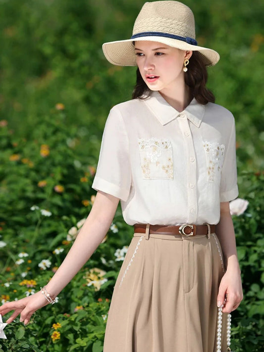 Woman in embroidered short-sleeve blouse and beige skirt with belt, wearing a straw hat outdoors.