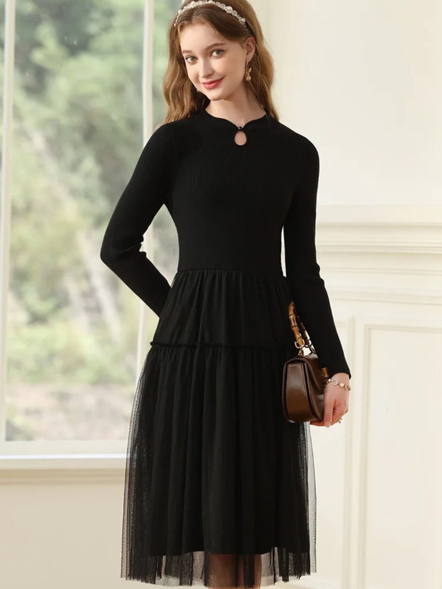 Woman in black long-sleeve dress with pleated mesh skirt and keyhole neckline, holding a brown handbag.