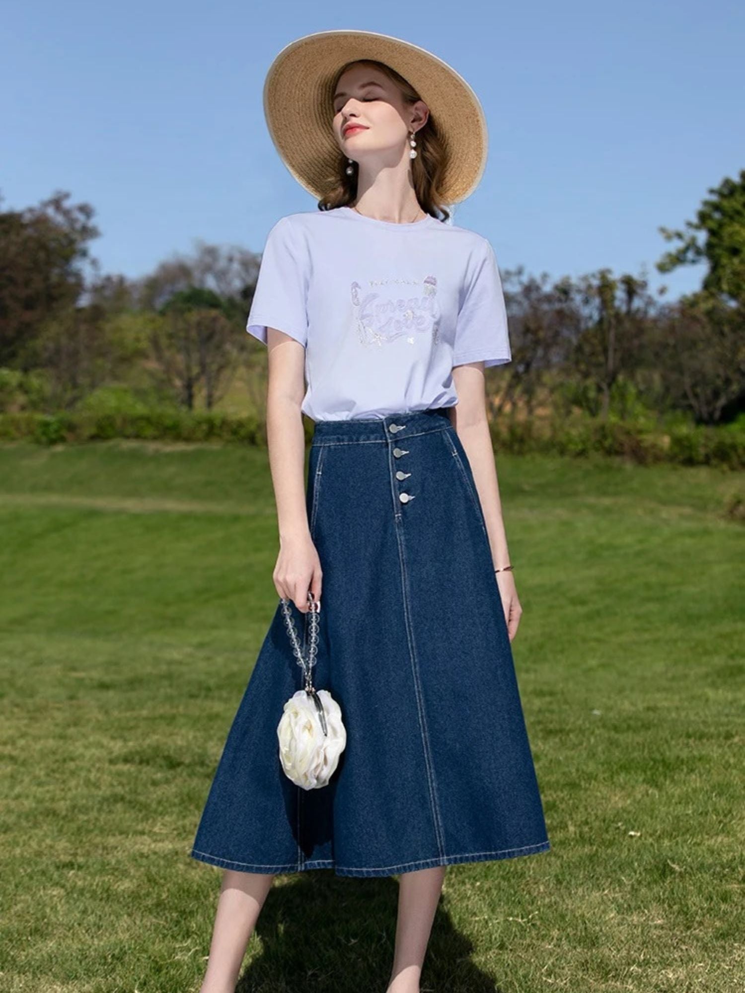 Woman in white graphic tee and A-line denim skirt with buttons, wearing a straw hat outdoors.