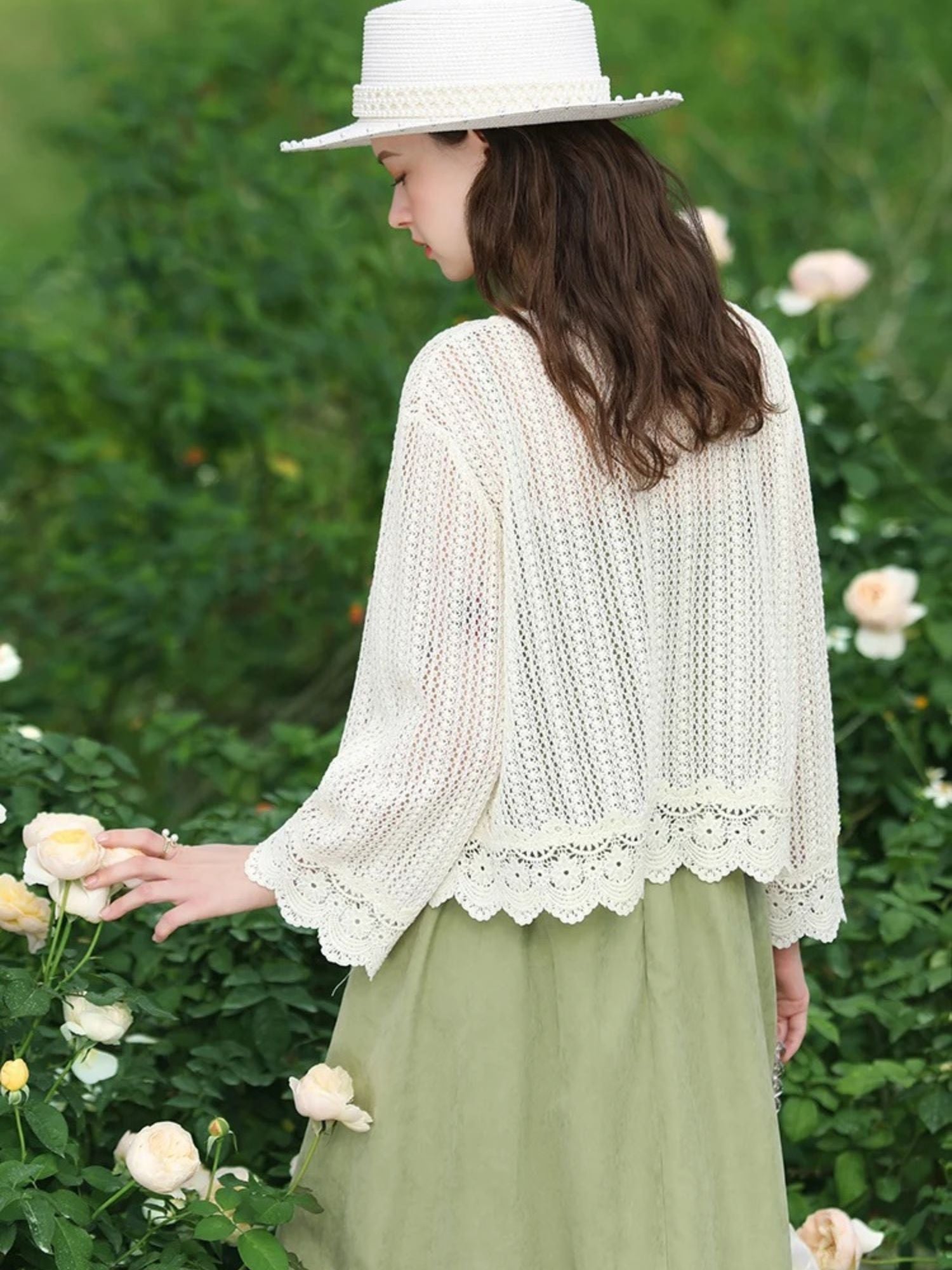 Back view of woman in white crochet cardigan and green dress, touching a blooming rose.