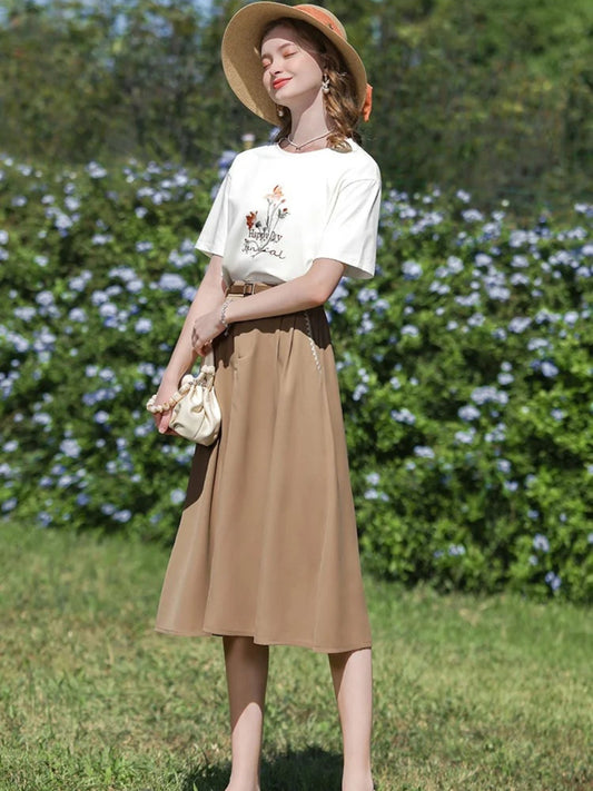 Woman in printed white tee and tan midi skirt smiling in a straw hat, holding a small handbag.