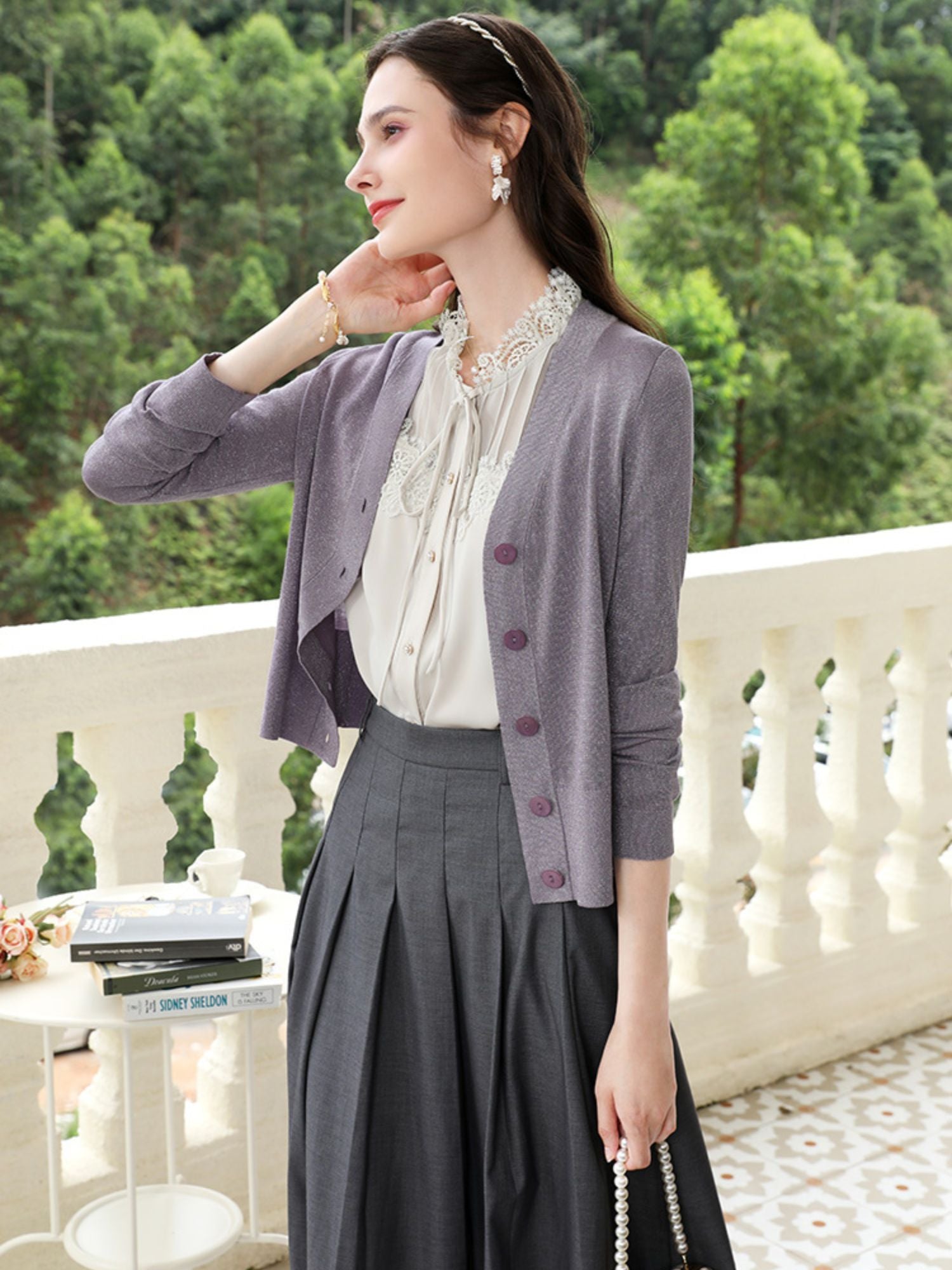 Woman in light purple cardigan and pleated skirt with ruffled blouse, touching her neck on a balcony.