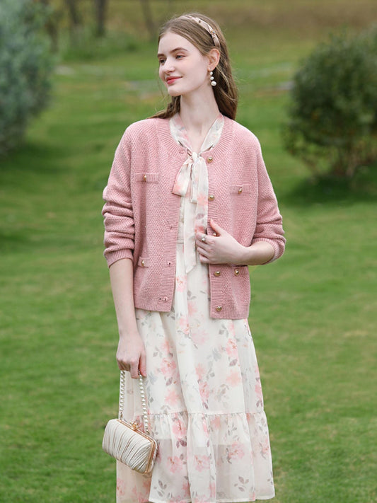 Woman in pink textured cardigan and floral dress holding a quilted purse, smiling softly outdoors.