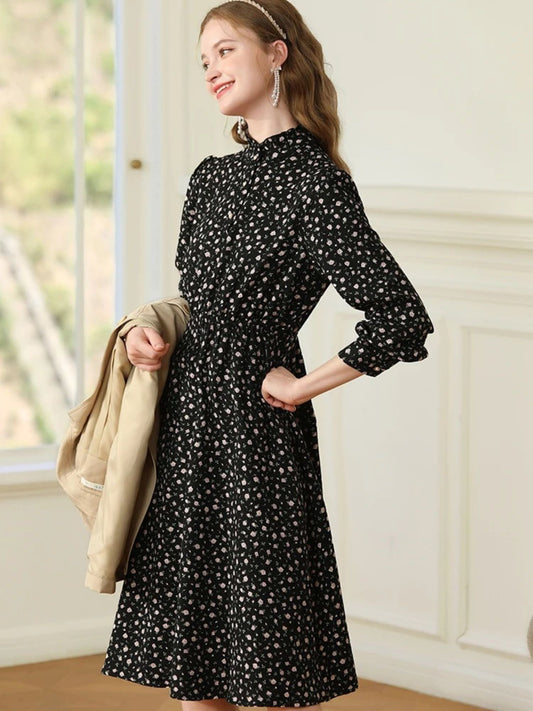 Woman in black floral dress with a gathered waist, holding a light coat in one hand.