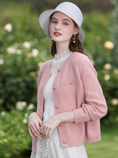 Woman in pink cardigan and white dress standing in a rose garden with a relaxed expression.

