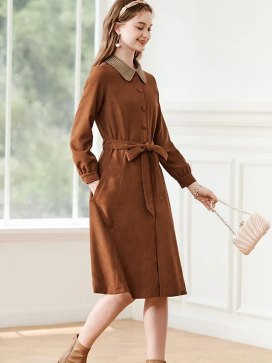 Woman walking in brown dress with tie waist and patterned collar, holding a beige clutch.