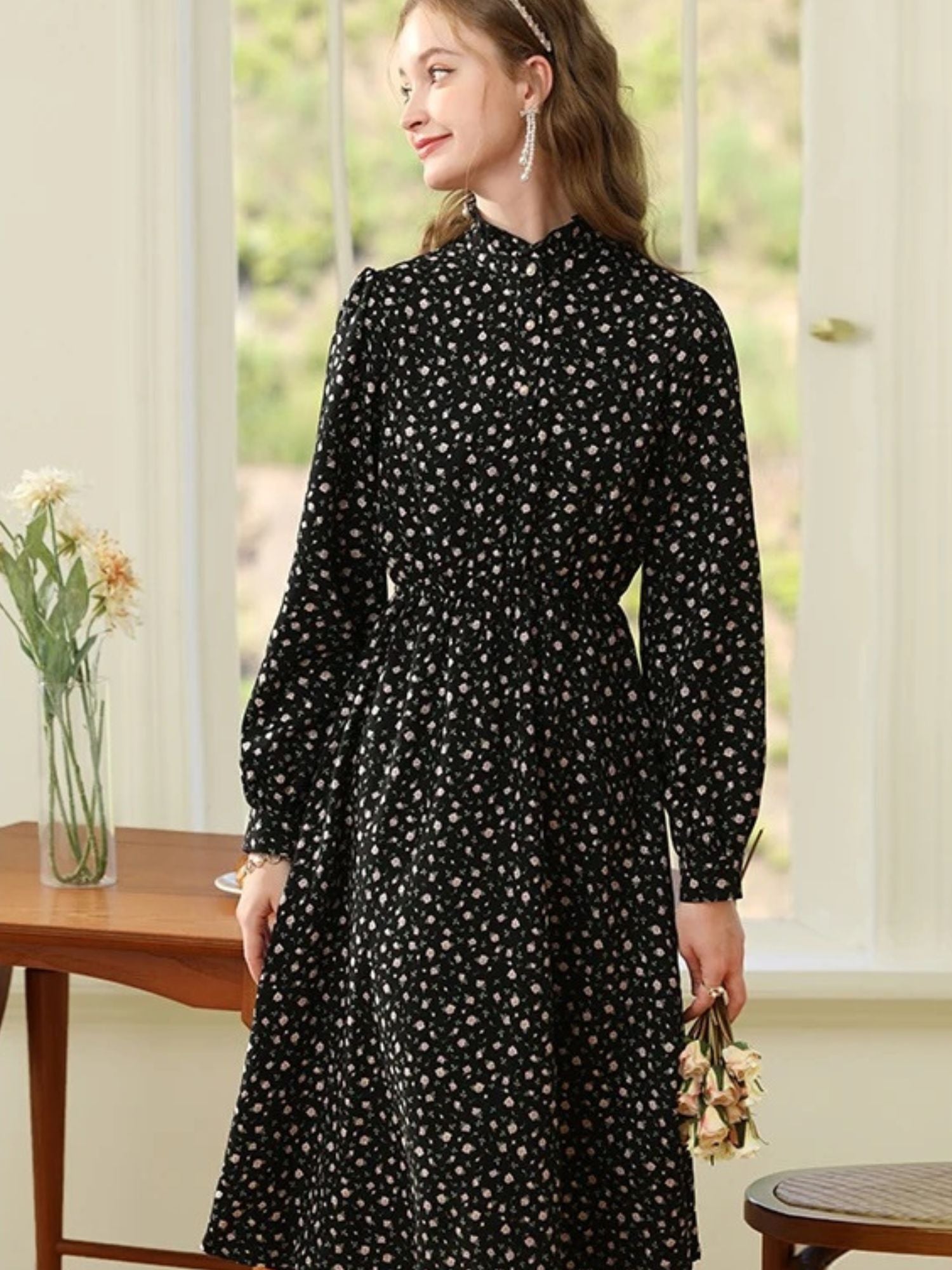 Woman in black and white floral dress standing near a wooden table with hands by her sides.