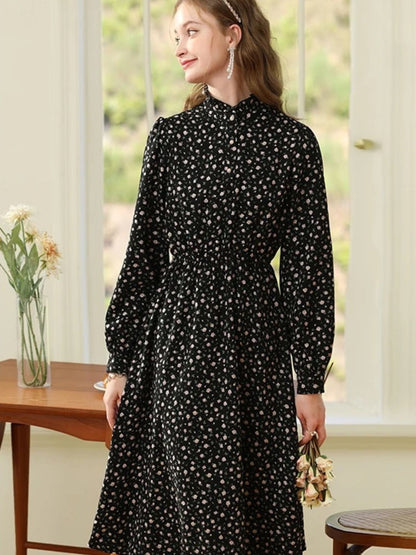 Woman in black and white floral dress standing near a wooden table with hands by her sides.