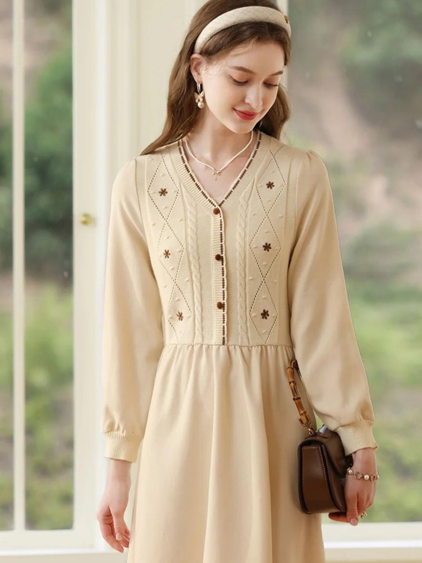 Woman in long-sleeve cream dress standing near a window with hands relaxed by her sides.