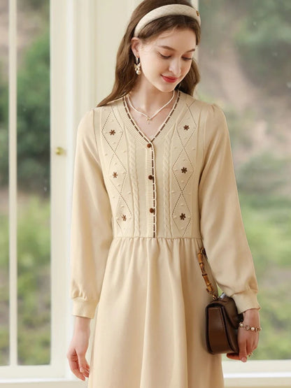 Woman in long-sleeve cream dress standing near a window with hands relaxed by her sides.