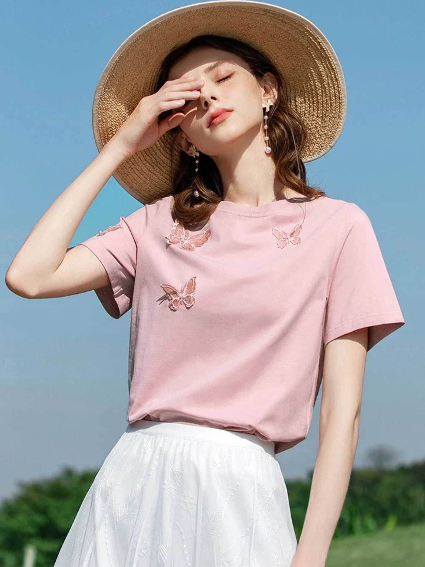 Woman shielding eyes from sun in pink butterfly top and white pleated skirt.