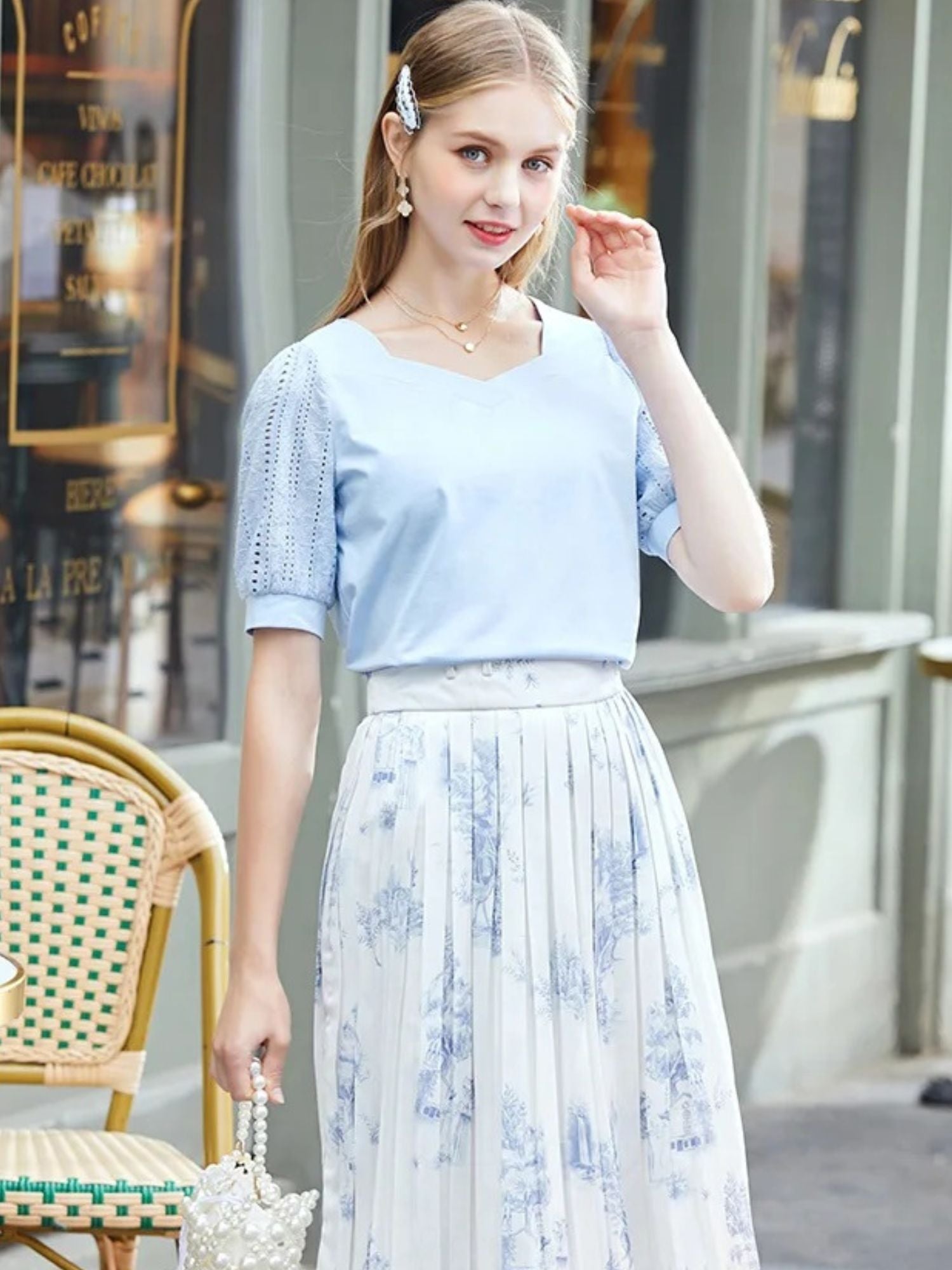 Woman standing in blue top and floral skirt, touching her cheek and smiling.