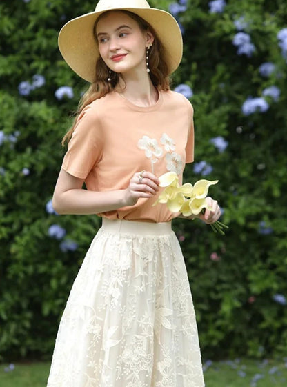 Woman in embroidered skirt and peach tee holding flowers, smiling near a floral hedge.