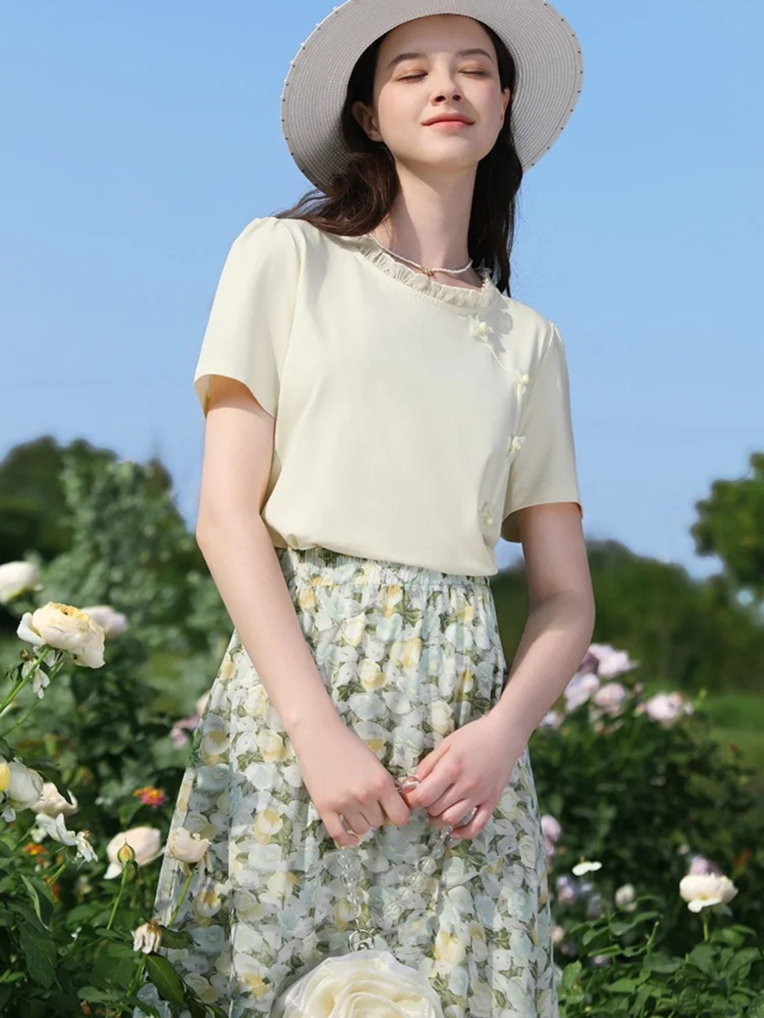 Woman in floral skirt and beige tee smiling under the sun, wearing a straw hat.