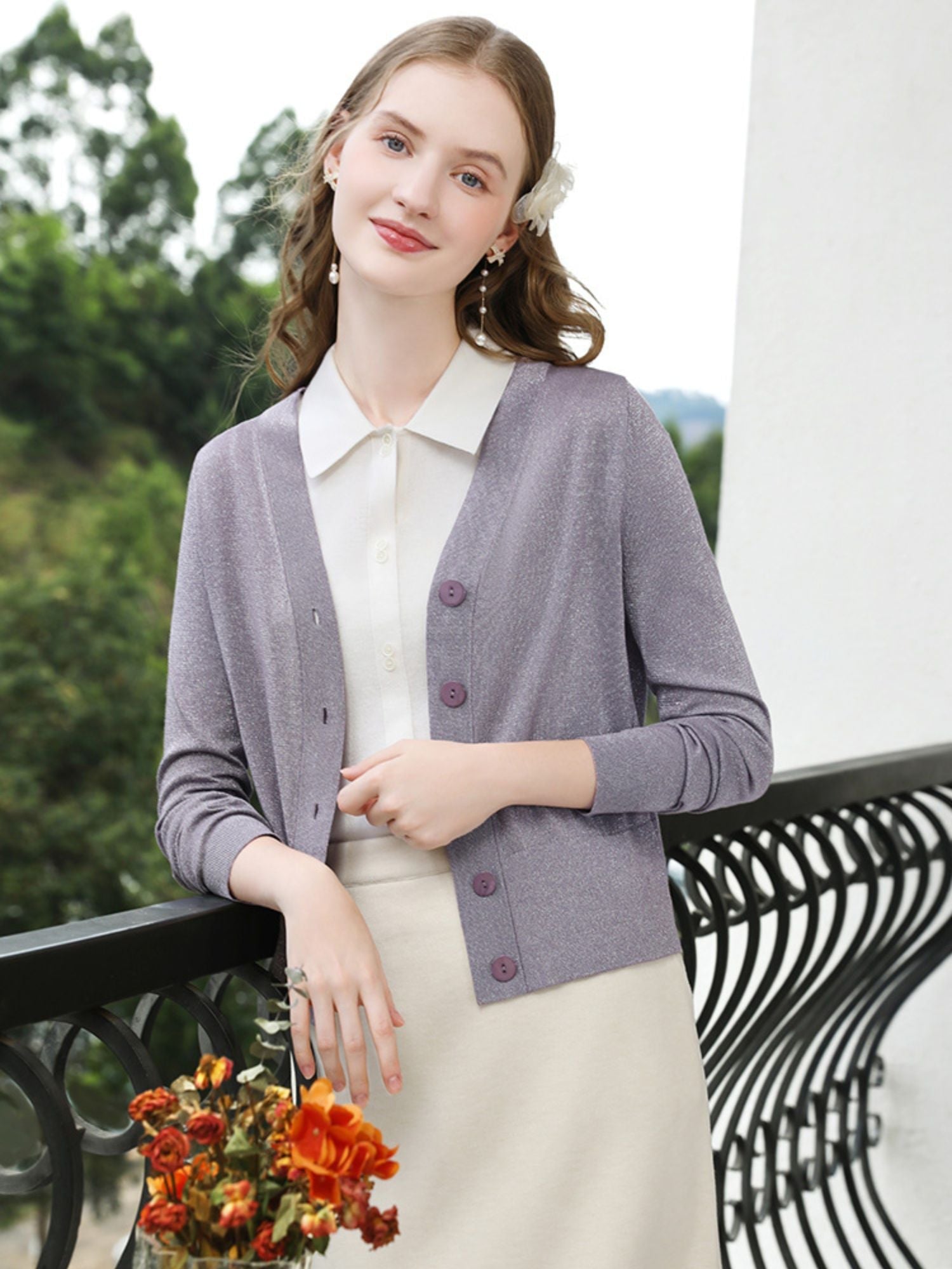 Woman smiling in purple cardigan and cream dress, holding a drink on a balcony.