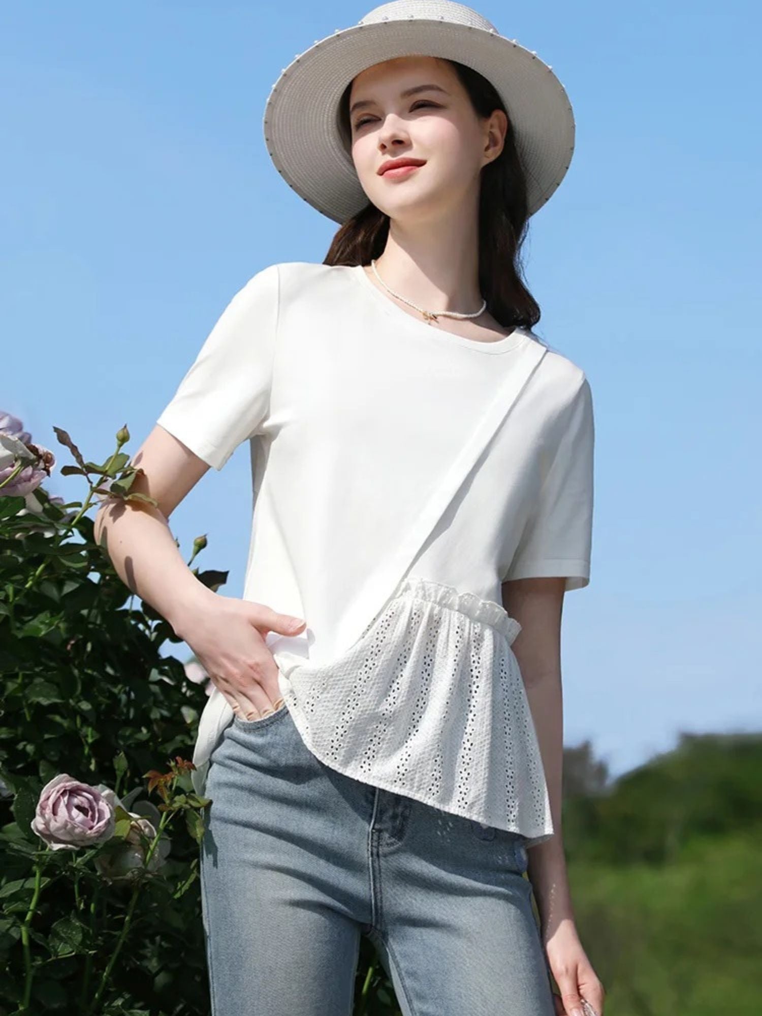 Woman in white short-sleeve top with lace hem, standing sideways near flowers, looking up.