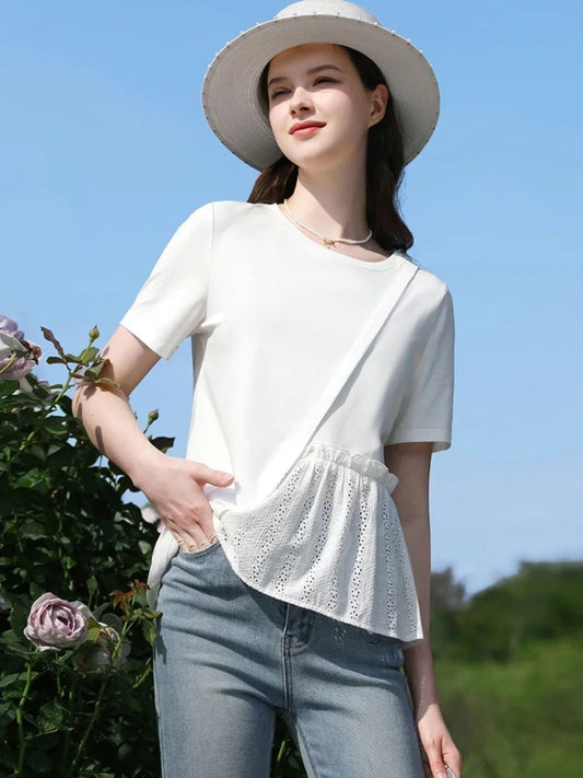 Woman in white short-sleeve top with lace hem, standing sideways near flowers, looking up.