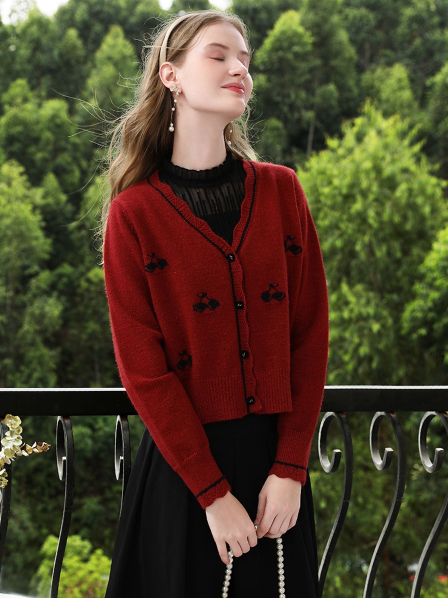 Woman in red floral knit cardigan smiling slightly, looking to the side on a balcony.