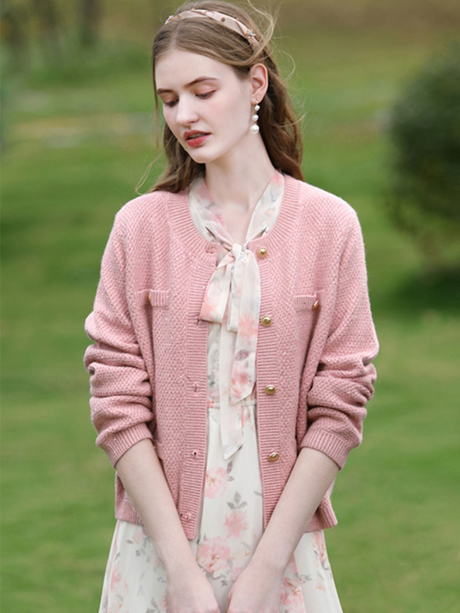 Woman in pink knit jacket and floral dress looking down with hands in pockets.