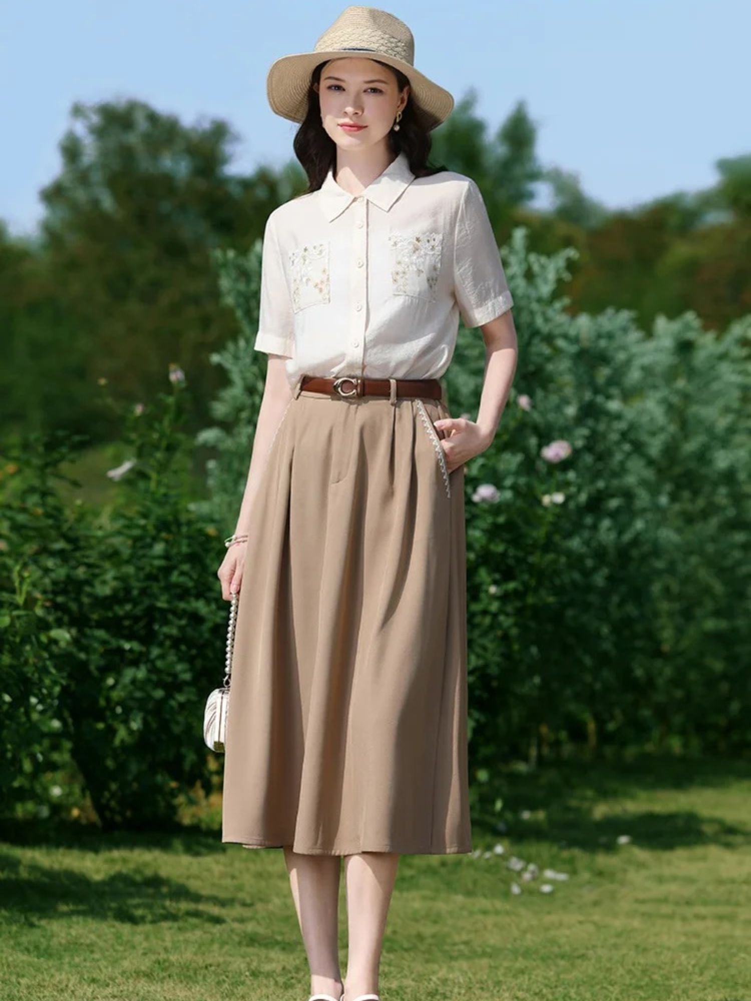 Woman standing in greenery wearing a short-sleeve blouse and midi skirt with a brown belt.