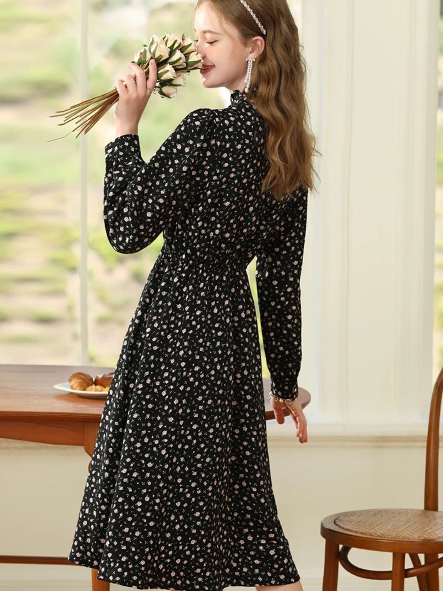 Woman in black floral midi dress holding a white flower and smiling by a chair.