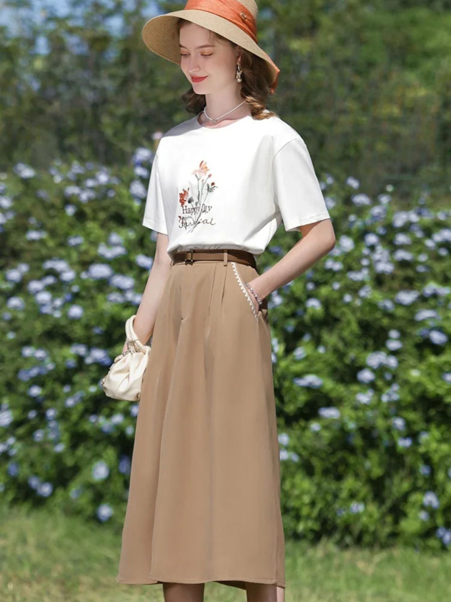 Woman in tan A-line skirt and white tee standing by a hedge of blue flowers.