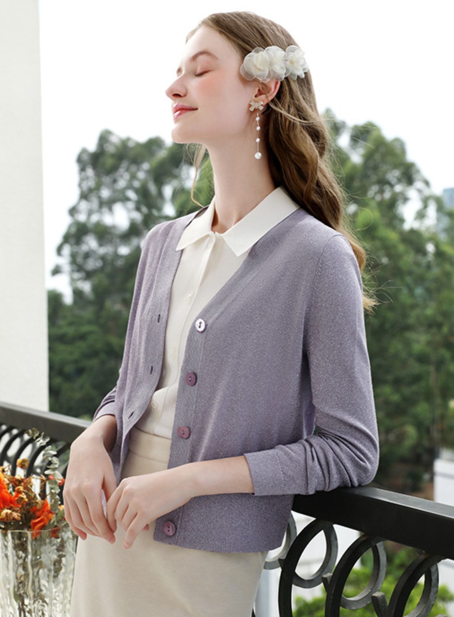 Woman in light purple sweater over pleated skirt looking upward near a floral centerpiece.