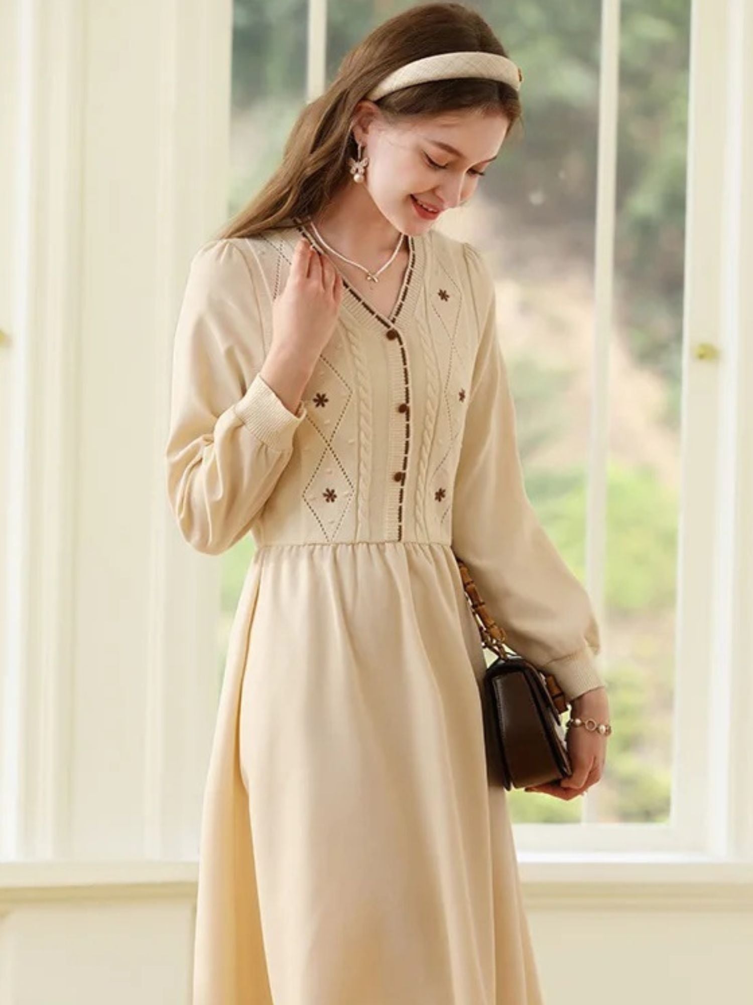Woman in beige dress looking down with one hand in her hair, holding a small purse.
