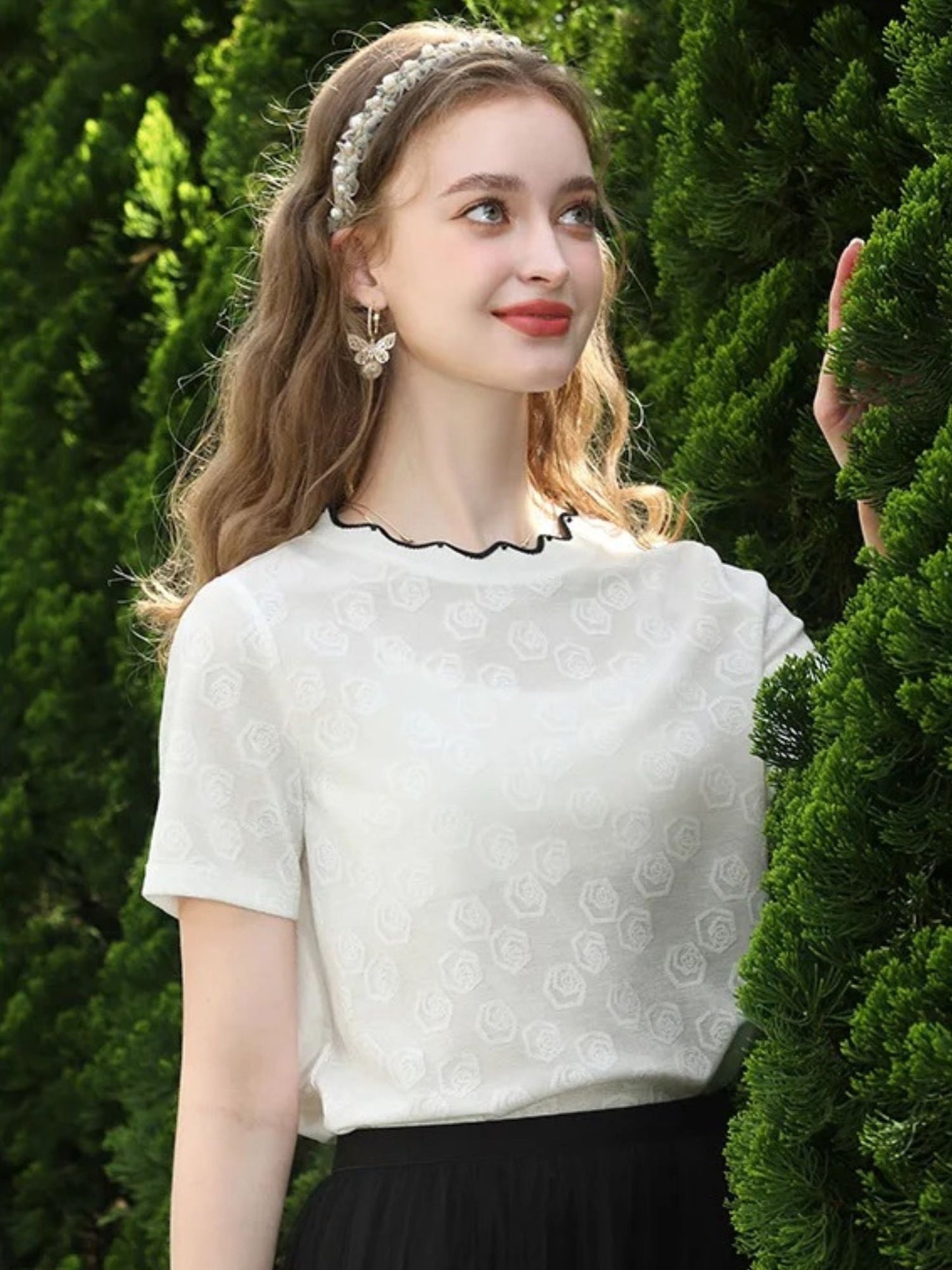 Woman in lace-patterned white top with scalloped neckline, standing in front of greenery.