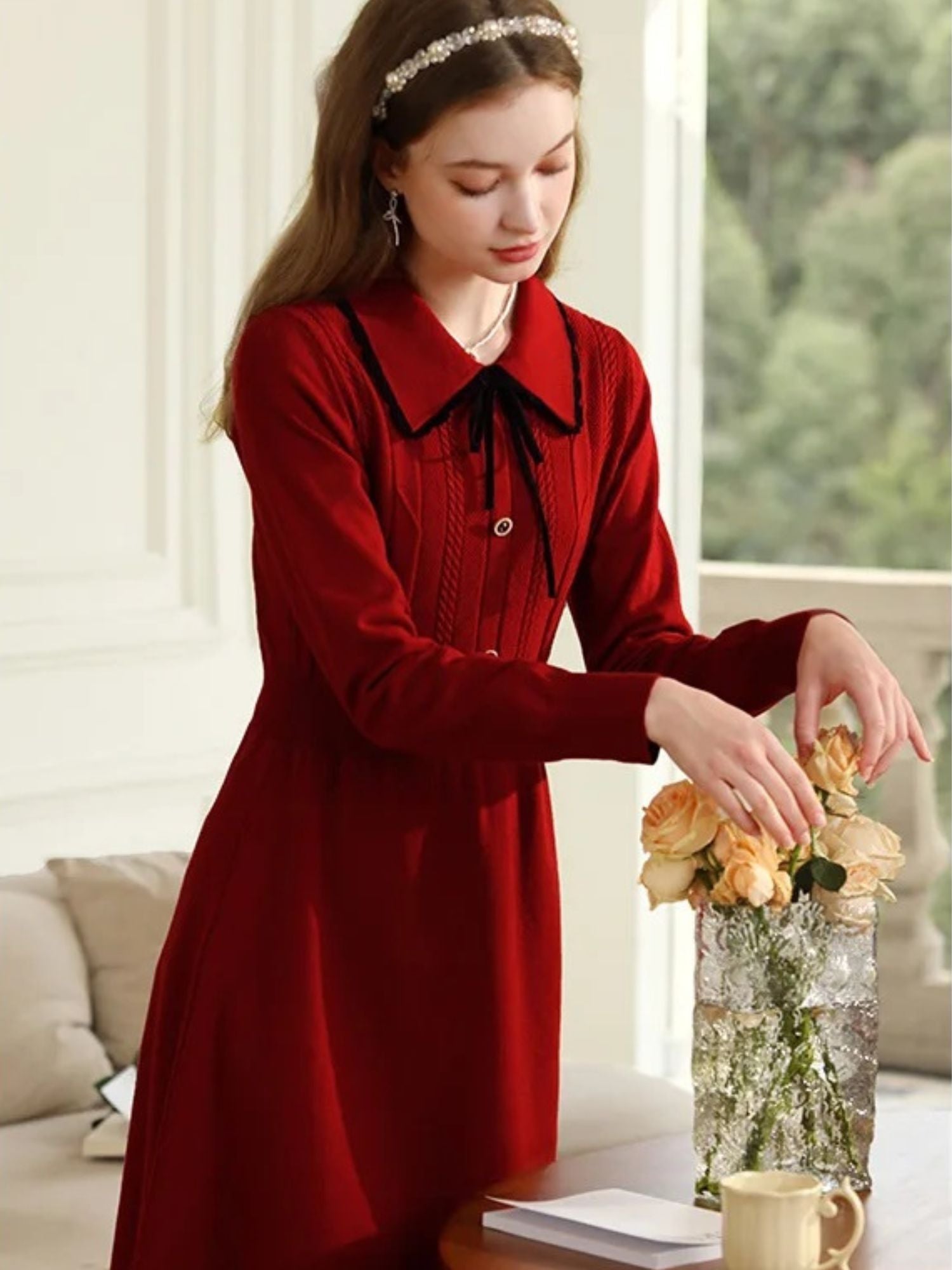 Woman in red dress holding flowers beside a white sofa, looking down.
