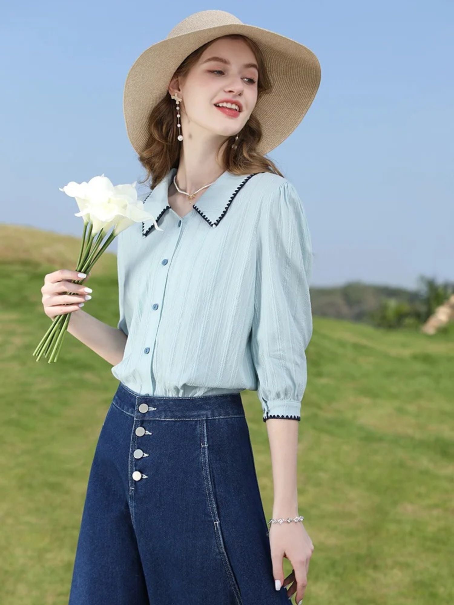 Woman in mint blouse and button-front denim skirt holding white flowers in a field.