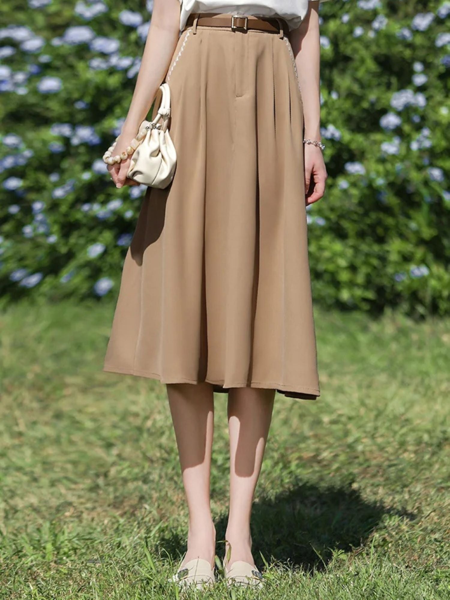 Front view of tan skirt with soft pleats, paired with a floral bag and sandals.